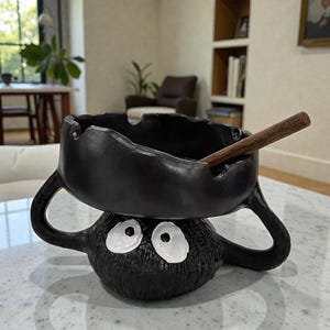May include: Black ceramic ashtray with a cartoon design. The ashtray has a bowl shape with a handle on each side, and is supported by a round base with white eyes. A wooden cigar rests in the bowl.