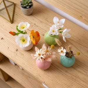 May include: Four small, round ceramic vases in white, light green, pink, and teal, each containing a different arrangement of artificial flowers and dried plants, including white and orange poppies, white blossoms, and small fluffy stems.