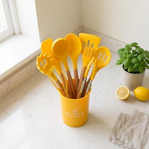 Op de afbeelding: Een set gele siliconen keukengerei met houten handvatten, waaronder een garde, soeplepel, spatel en tang, opgeborgen in een bijpassende gele cilindrische houder met de witte tekst KITCHEN UTENSIL SET op de voorzijde.