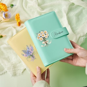 Könnte beinhalten: Zwei floral gestaltete Notizbücher werden in den Händen gehalten. Eines ist hellgelb mit einem Strauß blauer und violetter Blumen. Das andere ist mintgrün mit einem Gänseblümchenstrauß und dem Namen "Madison" auf dem Verschluss.