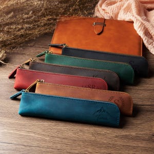 Op de afbeelding: Een collectie van zes slanke, rechthoekige etuis in verschillende kleuren, waaronder blauw, bruin, rood, donkerbruin, groen en zwart, elk voorzien van een metalen ritssluiting en een uniek gegraveerd berg- of bladontwerp op de voorzijde.