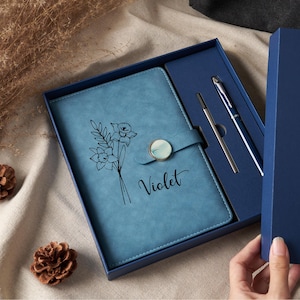 May include: A blue notebook with a floral line art illustration and the name Violet printed in black script. The notebook features a magnetic closure with a circular accent and is presented in a blue gift box with a matching blue pen and refill.