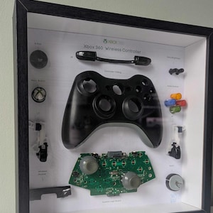 May include: A framed display showcasing the components of an Xbox 360 Wireless Controller. The black controller is central, with parts like buttons and circuit board arranged around it. The text "Xbox 360 Wireless Controller" is visible.
