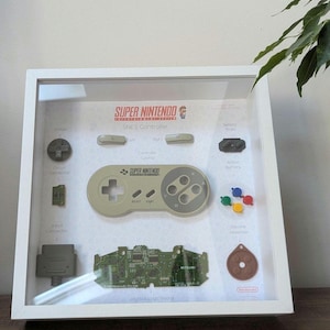 May include: A white framed display featuring a disassembled Super Nintendo controller. Components include the grey controller casing, D-pad, L and R shoulder buttons, colourful action buttons, a circuit board, input connector, and silicone absorber.