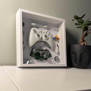 May include: White framed shadow box art featuring a disassembled Xbox 360 Wireless Controller. The controller's components are arranged inside, including the main body, circuit board, buttons, and joysticks. The text "Xbox 360 Wireless Controller" is visible.