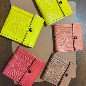 Könnte beinhalten: Eine Sammlung farbenfroher, geprägter Ledergeldbörsen. Die Geldbörsen sind in Gelb-, Rot-, Orange- und Brauntönen gehalten und weisen jeweils einzigartige Muster und einen Druckknopfverschluss auf. Die Geldbörsen sind auf einer braunen Oberfläche angeordnet.