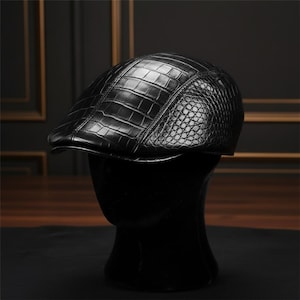 May include: A black leather newsboy cap with a textured, crocodile-like pattern. The cap is displayed on a black mannequin head, showcasing its shape and design. The hat has a curved brim and a rounded crown.
