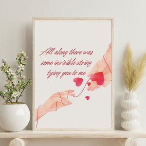 Puede incluir: Impresión artística enmarcada con el texto "All along there was some invisible string tying you to me." La obra muestra dos manos conectadas por un hilo rojo, con corazones rojos flotantes. La impresión está en una estantería de madera.