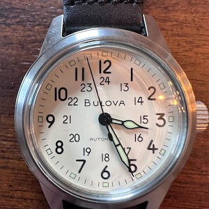 May include: A Bulova automatic wristwatch with a cream-colored face, black numerals, and luminous hands. The watch has a stainless steel case and a black leather strap. The watch face displays both 12-hour and 24-hour time.