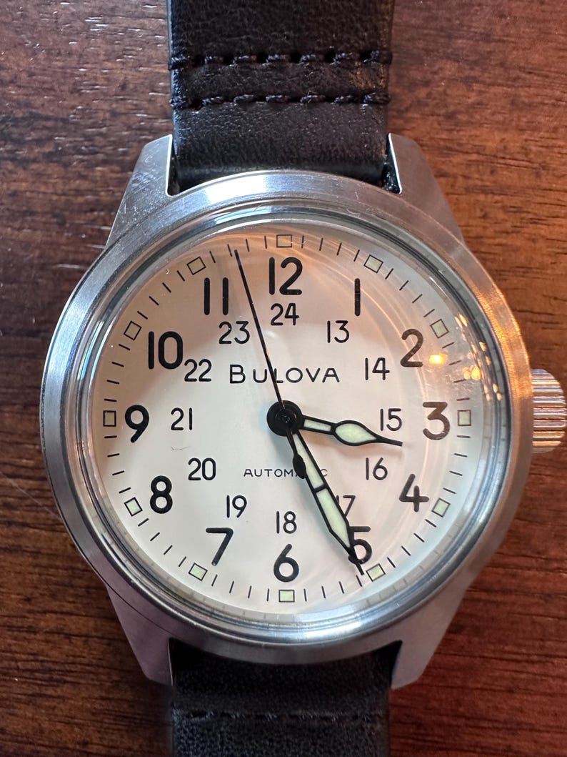 May include: A Bulova automatic wristwatch with a cream-colored face, black numerals, and luminous hands. The watch has a stainless steel case and a black leather strap. The watch face displays both 12-hour and 24-hour time.