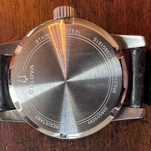 May include: A stainless steel Bulova watch with a black leather strap. The watch back is visible, displaying the brand name, model numbers, and the words "Water Resistant" and "Stainless Steel".