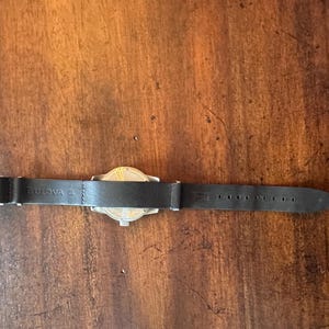 May include: A Bulova watch with a black leather band. The watch face is silver and gold. The band has a silver buckle and is marked with the Bulova brand name.