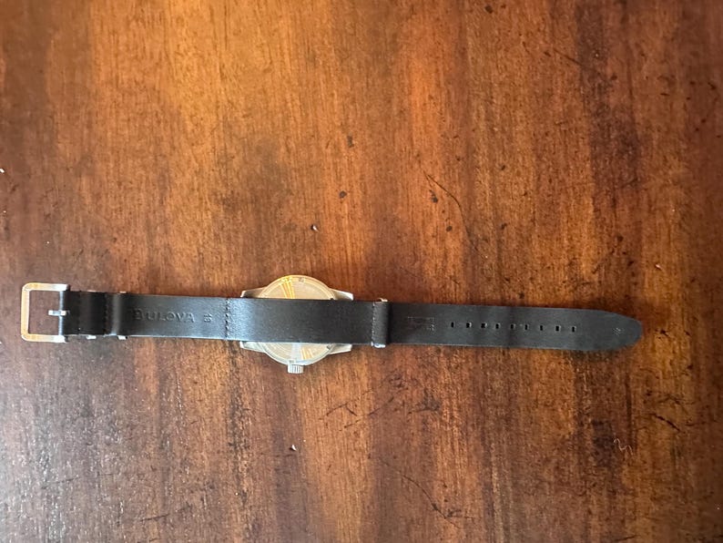 May include: A Bulova watch with a black leather band. The watch face is silver and gold. The band has a silver buckle and is marked with the Bulova brand name.