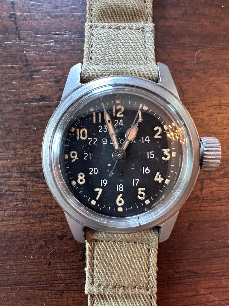 May include: A vintage wristwatch with a black face and a khaki canvas band. The watch has white numerals and hands, with a second set of numbers in a 24-hour format. The watch case is silver-colored.