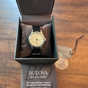 May include: A Bulova watch with a black leather band and a white face, displayed in its original box. The watch face features black numerals and hands. The box includes setting instructions and warranty information.