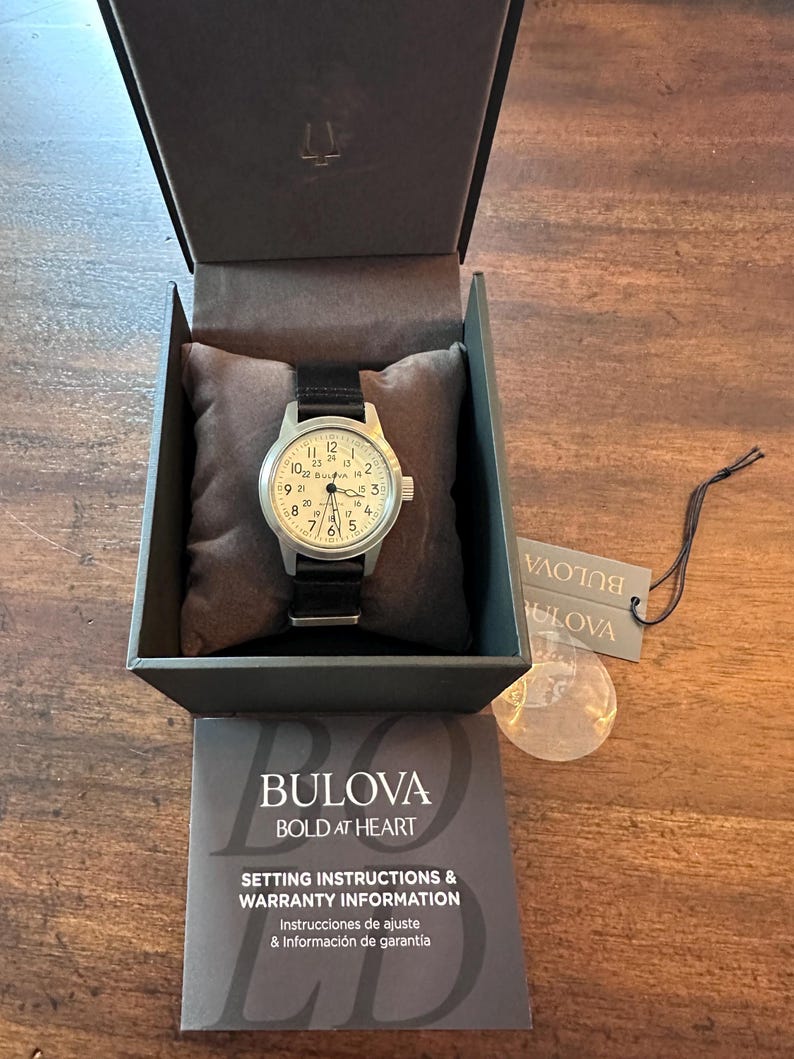 May include: A Bulova watch with a black leather band and a white face, displayed in its original box. The watch face features black numerals and hands. The box includes setting instructions and warranty information.