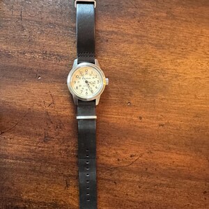 May include: A wristwatch with a black leather band and a silver-colored case. The watch face is off-white with black numerals and hands. The watch band is secured with a silver buckle.