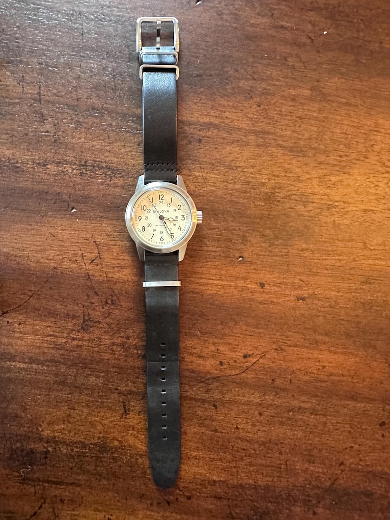 May include: A wristwatch with a black leather band and a silver-colored case. The watch face is off-white with black numerals and hands. The watch band is secured with a silver buckle.