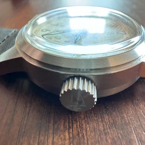 May include: Close-up of a silver-toned wristwatch with a black leather band. The watch has a clear, domed crystal and a textured crown. The watch face is visible through the crystal.