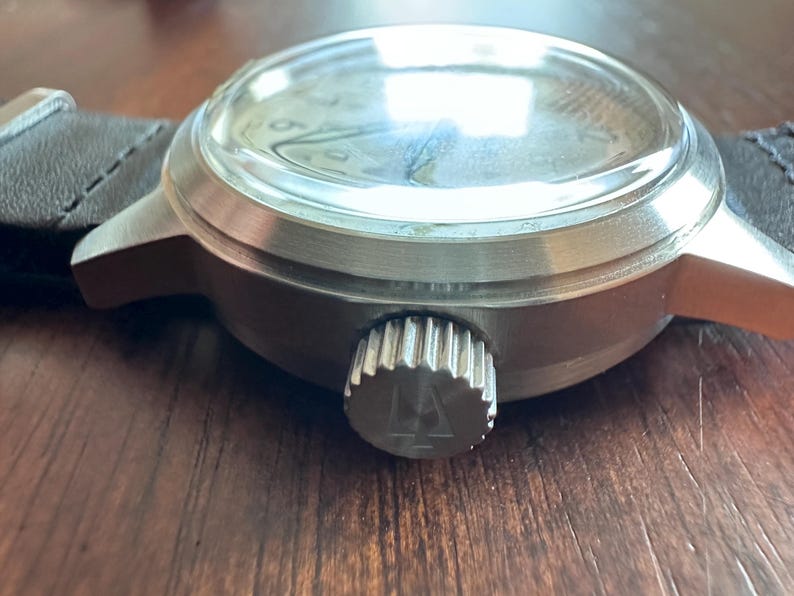 May include: Close-up of a silver-toned wristwatch with a black leather band. The watch has a clear, domed crystal and a textured crown. The watch face is visible through the crystal.
