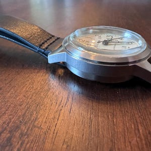 May include: A silver-toned wristwatch with a white face and black hands. The watch has a black leather band. The watch face displays numbers and markings. The watch is on a wooden surface.