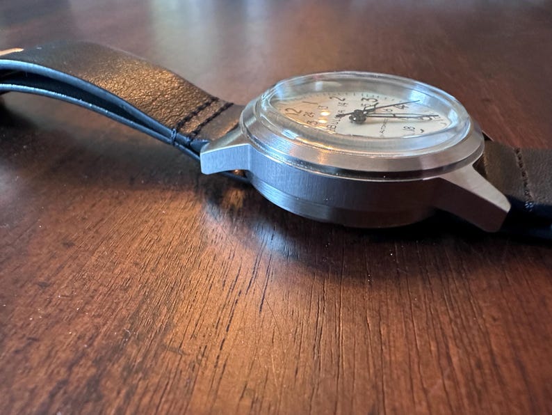May include: A silver-toned wristwatch with a white face and black hands. The watch has a black leather band. The watch face displays numbers and markings. The watch is on a wooden surface.