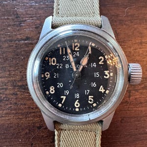 May include: A vintage wristwatch with a black face and a silver-colored case. The watch features white numerals and hands, with a second set of numbers in a 24-hour format. The watch has a khaki-colored canvas strap.