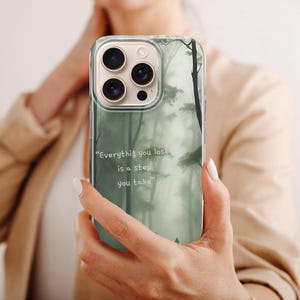 May include: A clear phone case featuring a misty forest design in shades of green and gray. The case has the quote, "Everything you lose is a step you take." The phone case is held up in front of a person.