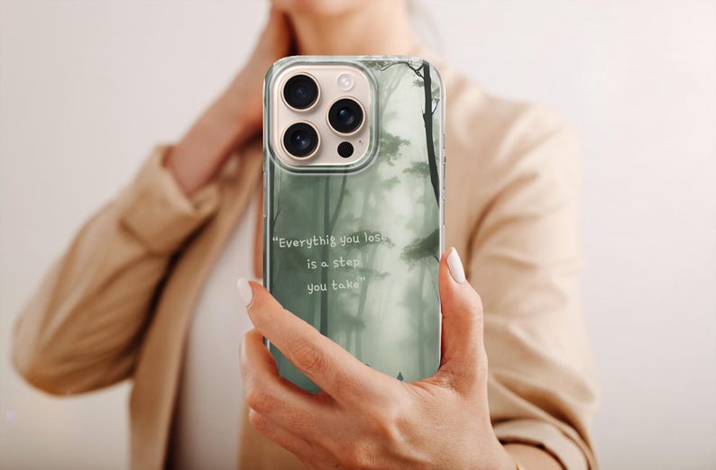 May include: A clear phone case featuring a misty forest design in shades of green and gray. The case has the quote, "Everything you lose is a step you take." The phone case is held up in front of a person.