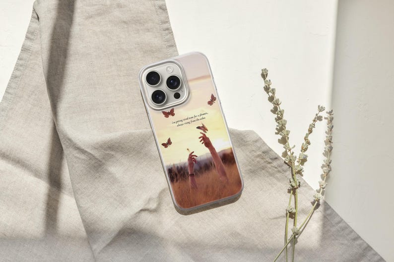 May include: A clear phone case with a nature-themed design. The case features a graphic of hands reaching for butterflies in a field, with text that reads "I'm going kind of crazy for a flower, always rising from the ashes."