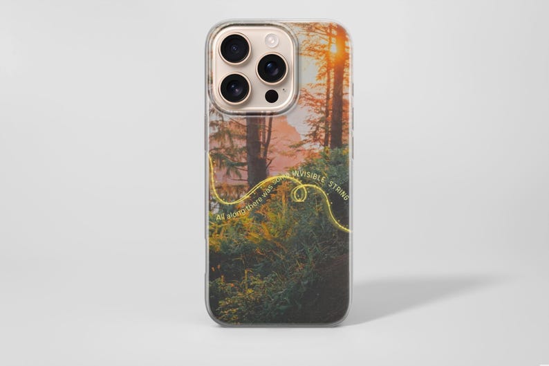 May include: Clear phone case featuring a forest scene with a sunset and the text "INVISIBLE STRING". The case has a gold-colored design element that follows the text "All along there was an". The phone case has a gold-colored camera ring.