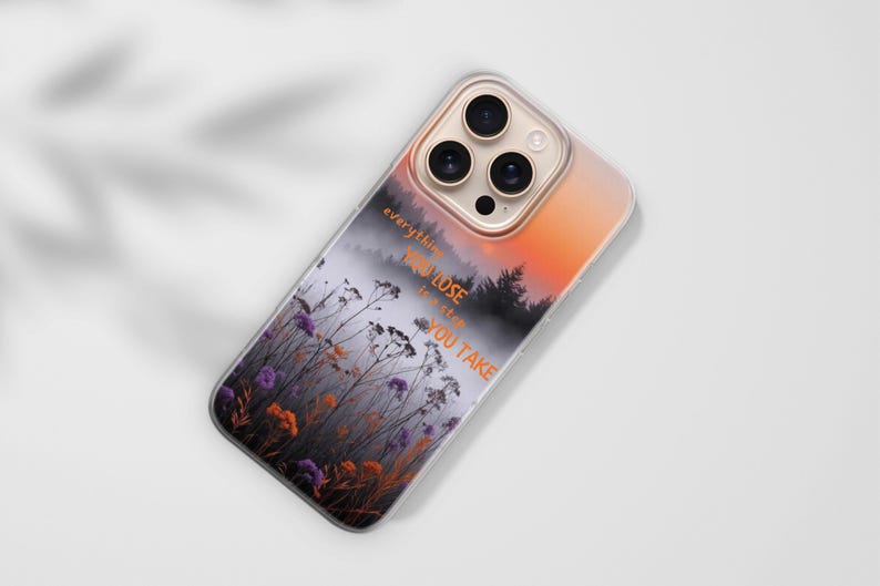 May include: A clear phone case with a landscape design featuring a misty forest, wildflowers, and an orange and pink sunset. The case has the quote "everything you lose is a step you take".