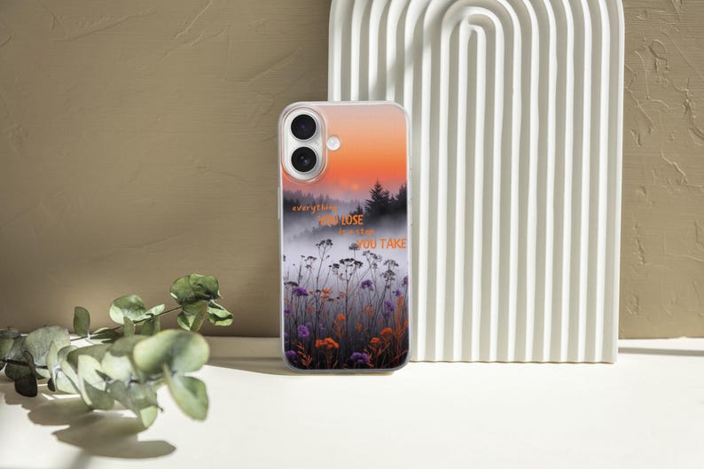 May include: A clear phone case featuring a sunset landscape with a forest silhouette and wildflowers. The case has the text "everything you lose is the chance you take" in orange. The case is on a white surface.