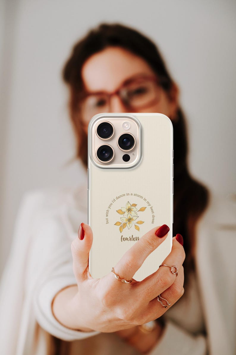 May include: A cream-colored phone case with a floral design and the word "fearless" in a script font. The case has a clear border and a camera cutout. The design includes the text "but with you I'd dance in a storm in my best dress."