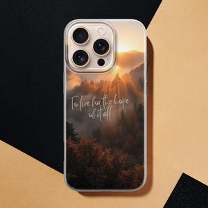 May include: Clear phone case featuring a scenic landscape with a forest and a sunset. The case has the text "To live for the hope of it all" in a cursive font. The phone case is on a black and tan surface.