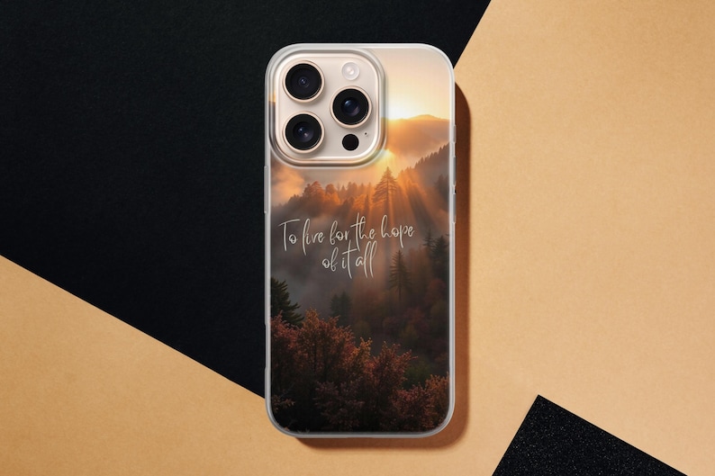 May include: Clear phone case featuring a scenic landscape with a forest and a sunset. The case has the text "To live for the hope of it all" in a cursive font. The phone case is on a black and tan surface.