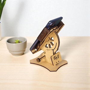 May include: A wooden phone stand with a dark phone in place. The stand is made of light-coloured wood and features a unique gear and cutout design. The base has a small wooden sign that says "Tree D".