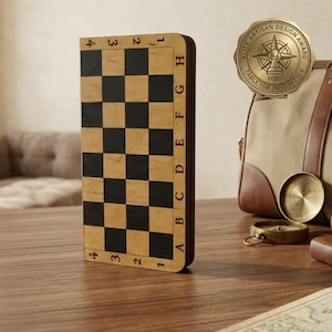 May include: A wooden chess board with black and tan squares, standing upright. The board has numbered and lettered edges. A gold-colored medallion with the text "Elite Artisan Design Award Tabletop Innovation 2026" is visible in the background.