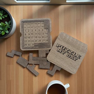 Op de afbeelding: Houten puzzelkalender met de tekst "PUZZLE MY DAY". De kalender heeft maanden en data, met individuele stukken voor de puzzel. De stukken zijn verspreid over een houten oppervlak.