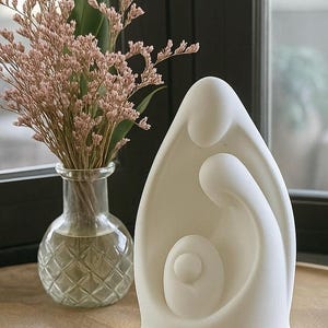 Può includere: Una scultura in ceramica bianca raffigurante una famiglia, con madre, padre e figlio. La scultura è su una superficie di legno, accanto a un vaso di vetro con fiori secchi rosa e foglie verdi. Lo sfondo comprende una finestra e modanature scure.