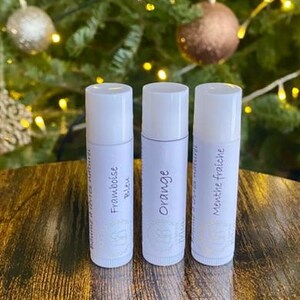 Peut inclure: Trois tubes de baume à lèvres blancs avec des étiquettes argentées, chacun avec un nom de saveur différent : "Framboise Bleu", "Orange" et "Menthe Fraiche". L'arrière-plan présente un sapin de Noël flou.