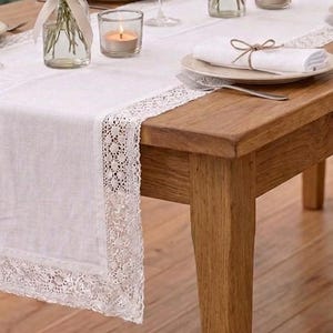 Puede incluir: Un camino de mesa de lino blanco con delicado encaje, colocado sobre una mesa de madera. El camino de mesa está adornado con velas, un jarrón con flores y cubiertos, creando una escena de comedor elegante.