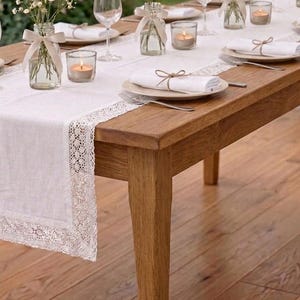 Puede incluir: Una mesa de comedor de madera preparada para una ocasión especial. La mesa está adornada con un camino de mesa blanco con encaje, platos blancos, servilletas y pequeños jarrones de cristal con flores. Velas encendidas y copas de vino completan el elegante ambiente.