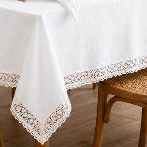 Peut inclure: Nappe en lin blanc avec une bordure en dentelle délicate. La nappe est drapée sur une chaise en bois, mettant en valeur la texture et le design. La bordure en dentelle ajoute une touche d'élégance. Parfait pour une salle à manger ou une cuisine.