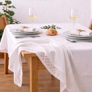 Puede incluir: Un mantel de lino blanco con un delicado ribete de encaje, sobre una mesa de madera. La mesa está puesta para una comida, con platos, cubiertos, servilletas, pan y copas de vino llenas de una bebida clara.