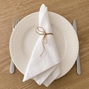 Puede incluir: Un plato de cerámica blanca con un patrón de hojas texturizado en el borde, que presenta una servilleta de tela blanca doblada y atada con un lazo de cuerda marrón. Un tenedor de plata está a la izquierda del plato y un cuchillo a la derecha.