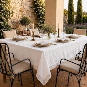 Puede incluir: Una mesa de comedor al aire libre preparada para una comida. La mesa está cubierta con un mantel blanco y está puesta con platos, cubiertos y vasos. Sillas de metal negro con cojines beige rodean la mesa. Velas y flores.