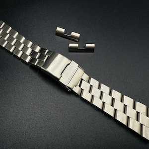 Könnte beinhalten: Ein silberfarbenes Metall-Uhrenarmband mit Faltschließe, bestehend aus rechteckigen Gliedern. Zwei zusätzliche Glieder befinden sich oberhalb des Armbands. Das Uhrenarmband wird vor einem dunklen Hintergrund gezeigt.