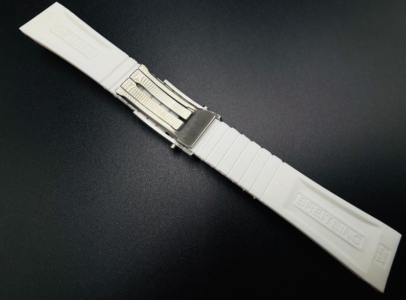 Puede incluir: Correa de reloj Breitling blanca con cierre plateado. La correa est&aacute; hecha de un material acanalado y texturizado. El logotipo de Breitling est&aacute; grabado en la correa.