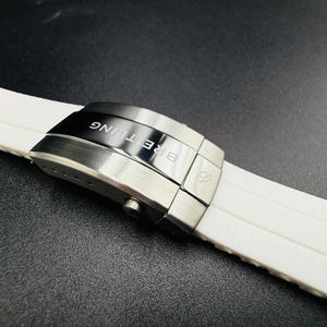 Puede incluir: Primer plano de una correa de reloj blanca con un cierre plateado. El cierre est&aacute; grabado con la palabra "BREITLING" en negro. La correa est&aacute; hecha de un material suave y flexible.